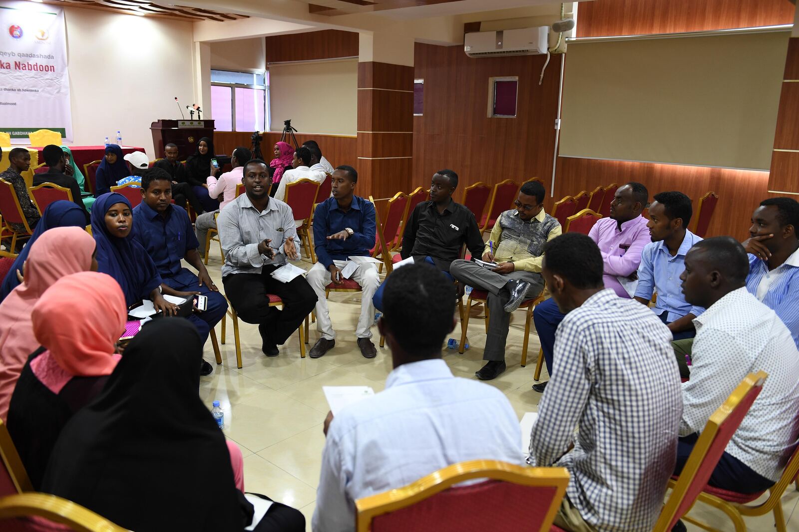 A large group sitting in a circle of chairs, one person speaking and the others listening