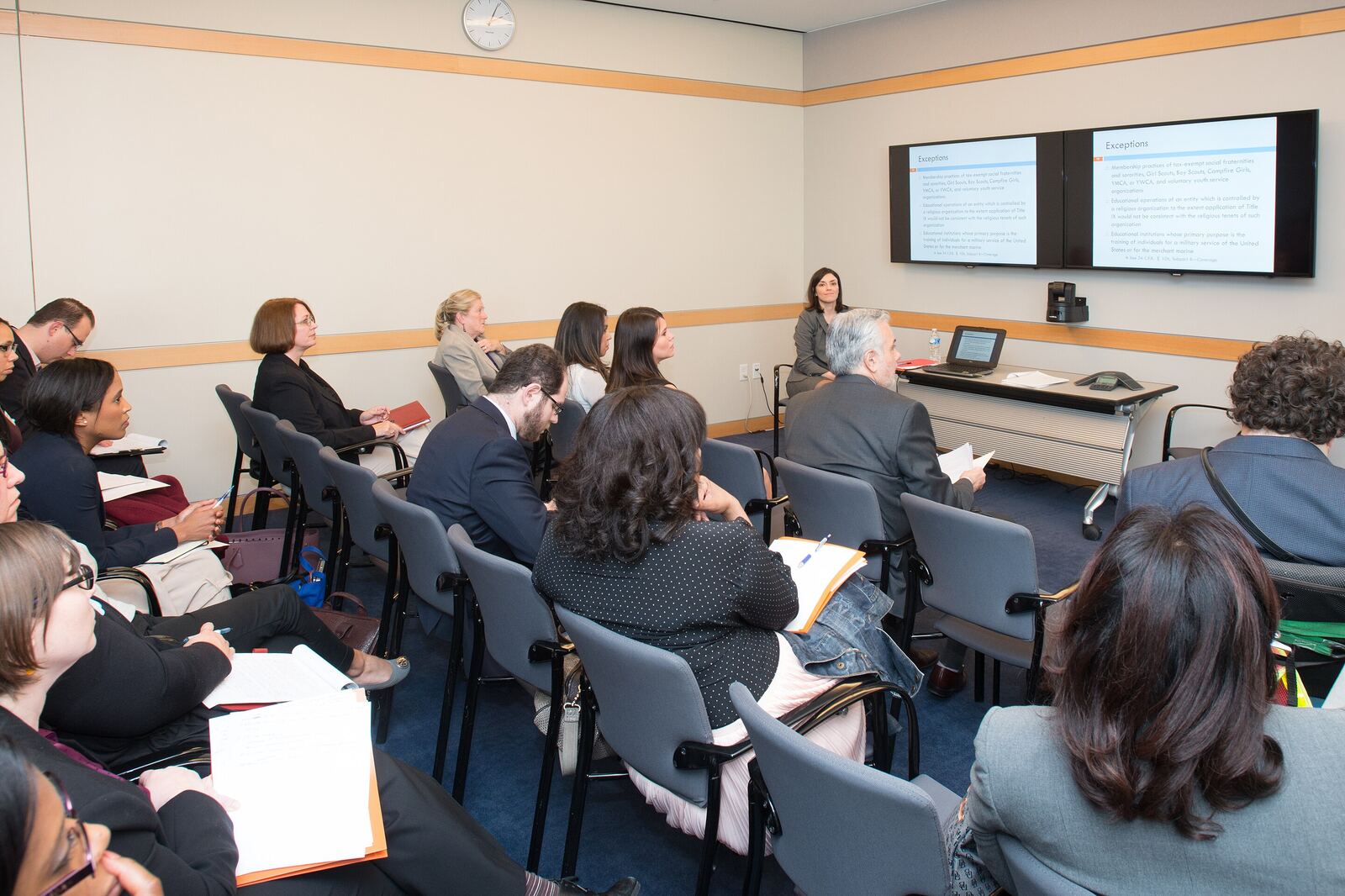 A meeting room where seated participants face a facilitator standing beside a screen